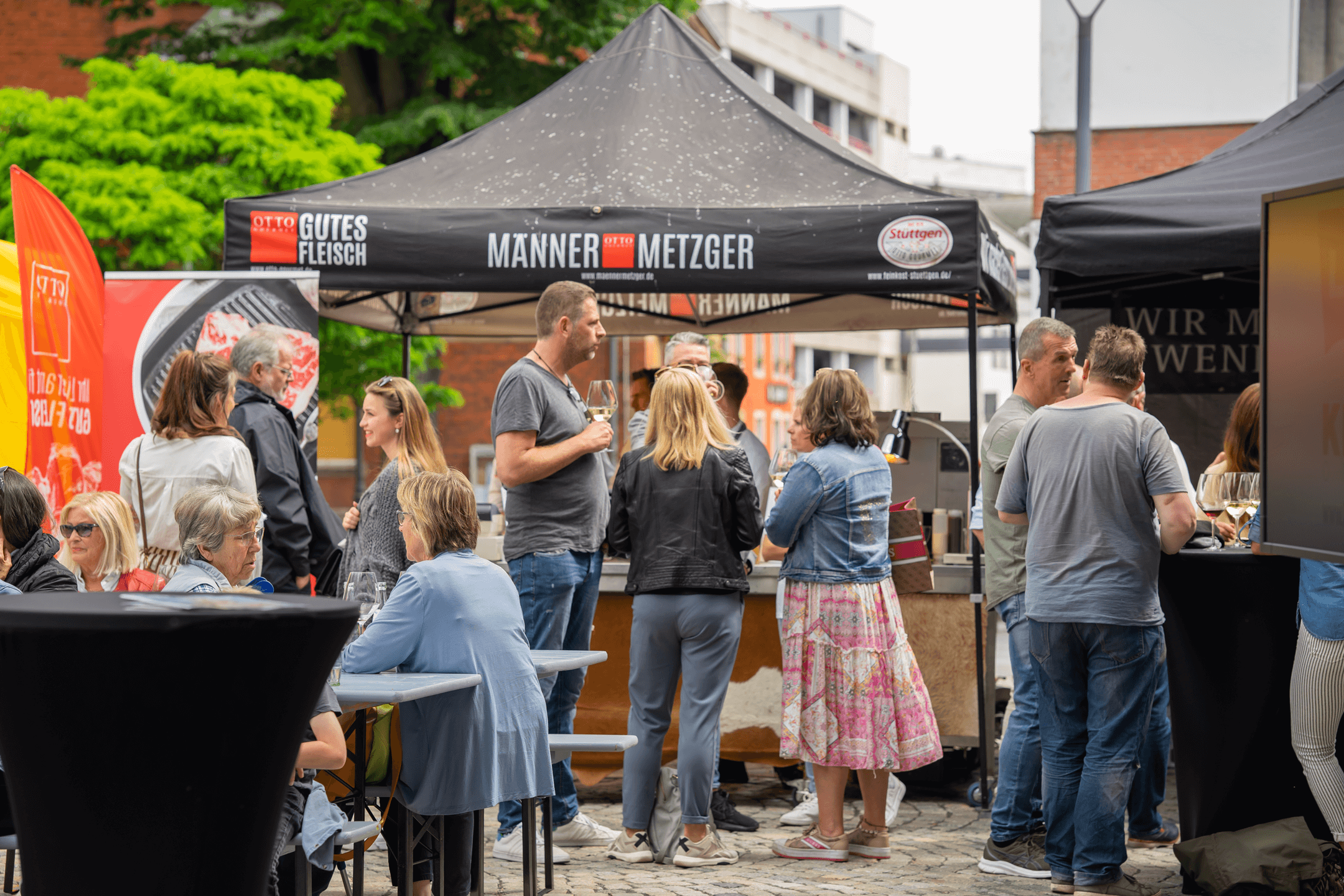 Eine Gruppe von Menschen steht vor einem Zelt der Männer Metzger by Otto Gourmet und genießt Speisen und Getränke während des 2. Mönchengladbacher Fashion Days, vor der Markthalle Mönchengladbachs.