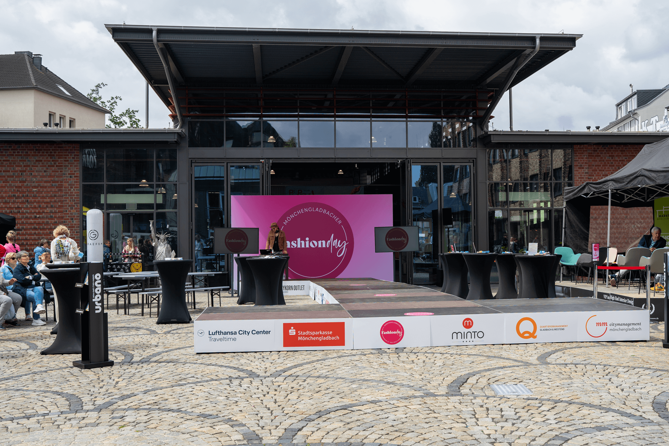 Aufnahme des aufgebauten Laufstegs für den 2. Mönchengladbacher Fashion Day vor der Markthalle Mönchengladbach. Um den Laufsteg herum stehen Stehtische mit schwarzen Hussen.