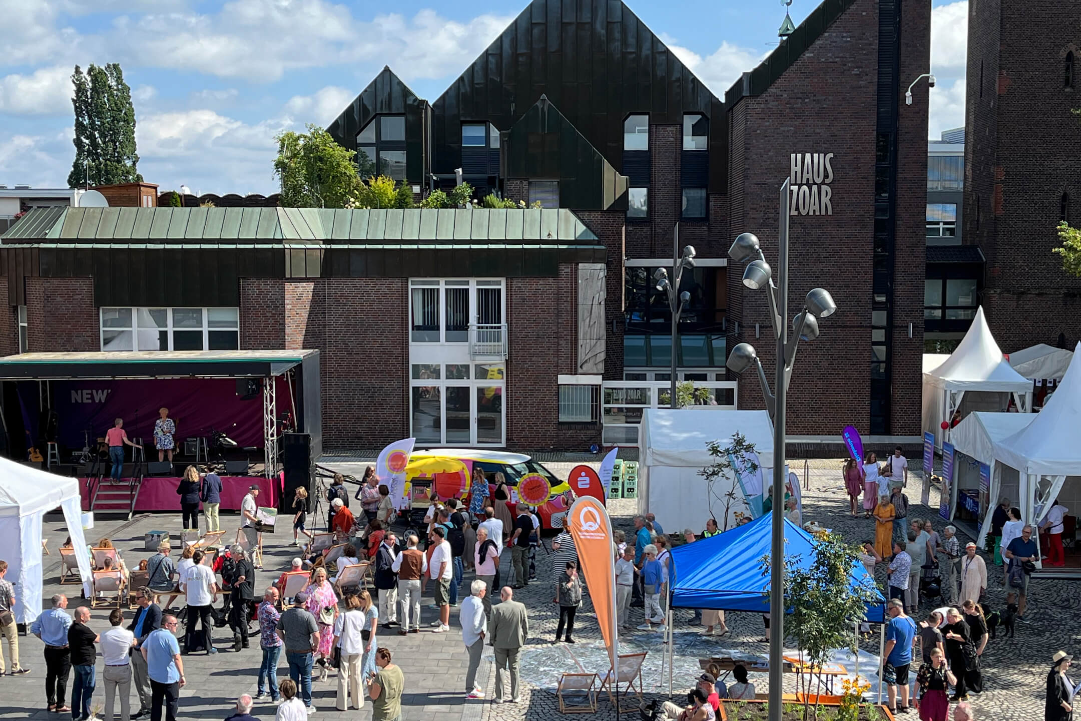 Bei gutem Wetter steht eine große Gruppe von Menschen versammelt um aufgebaute Zelte und Pavillons auf dem neu eröffneten Kapuzinerplatz, vor dem Haus Zoar.
